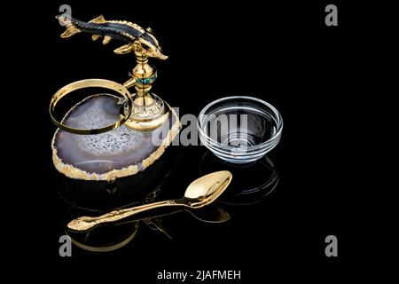 Gilded luxury crystal caviar bowl on stand made of natural quartz, carved brass fish figurine and dessert spoon on a black background. Stock Photo