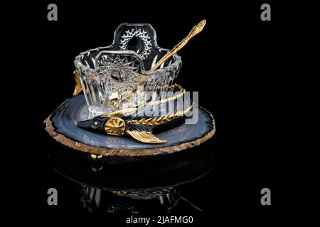 Gilded luxury crystal caviar bowl on stand made of natural quartz, carved brass fish figurine and dessert spoon on a black background. Stock Photo