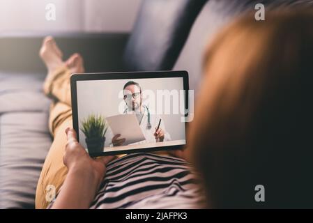 Woman lying on a sofa and talking with a doctor online using digital tablet. Telemedicine concept. Stock Photo