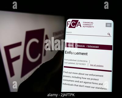 Person holding cellphone with webpage of British Financial Conduct Authority (FCA) on screen in front of logo. Focus on center of phone display. Stock Photo