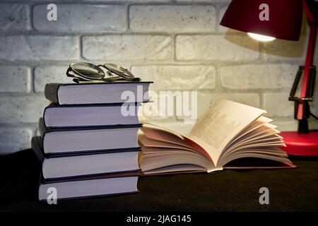 Books, glasses and a table lamp on a wall background. A table lamp illuminates an open book Stock Photo