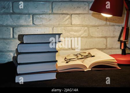 Books, glasses and a table lamp on a wall background. A table lamp illuminates an open book Stock Photo
