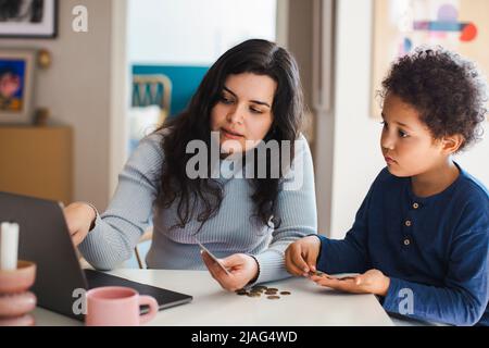 Son counting coins while sitting with mother using laptop at home Stock Photo