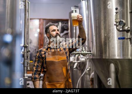 Bearded brewery master holding glass of beer and evaluating its visual characteristics. Small family business, production of craft beer. Stock Photo