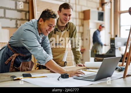 Warm toned portrait of animated carpenters using laptop and discussing furniture designs in workshop Stock Photo