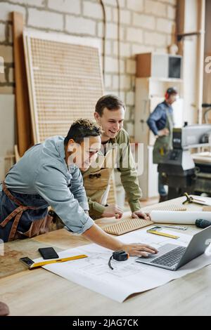 Vertical warm toned shot of two modern carpenters using laptop in workshop and discussing designs Stock Photo