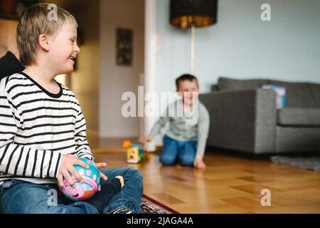 Happy boy having down syndrome with ball sitting on floor in living room Stock Photo