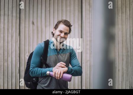 Portrait of happy young man with backpack and water bottle standing against wall Stock Photo