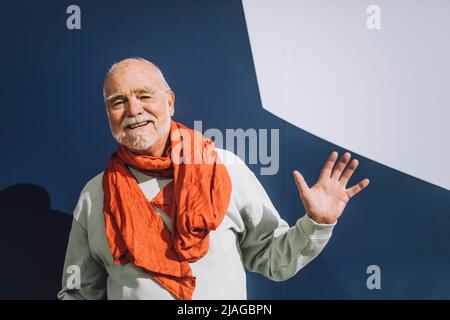 Portrait of happy senior man wearing scarf waving hand against blue and white wall Stock Photo