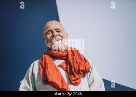 Portrait of cheerful senior man wearing orange scarf standing against blue and white wall Stock Photo