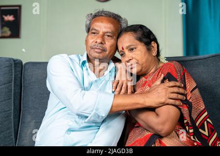 Senior man consoles by embracing his thouhgtful wife at home while on sitting sofa - concept of family, emotional love and support Stock Photo