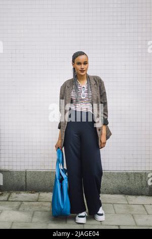 Full length portrait of young woman with bag standing on footpath Stock Photo