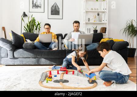 Parents work remotely with laptops sitting on the couch in living room, two sweet kids playing with toys on the foreground, freelancer parents working on the distance during quarantine Stock Photo