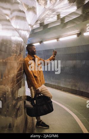 Full length of young man carrying bag while taking selfie through mobile phone in subway tunnel Stock Photo