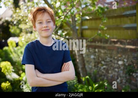 Portrait Of Smiling Boy With Crossed Arms Standing In Summer Garden At Home Stock Photo
