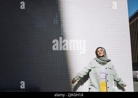 Smiling woman enjoying listening music through wireless headphones against wall Stock Photo