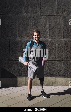 Portrait of smiling man with disability holding water bottle and exercise mat on footpath during sunny day Stock Photo