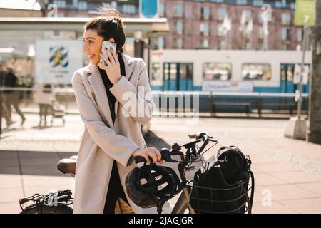 Businesswoman talking on smart phone standing with bicycle while looking back over shoulder in city Stock Photo