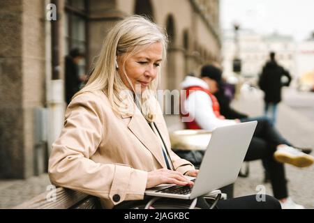 Blond businesswoman using laptop while sitting on bench in city Stock Photo