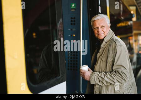 Side view of happy businessman holding disposable cup while boarding in train at railroad station Stock Photo