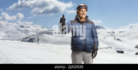 Mature man in a winter jacket standing in a ski resort on a snowy mountain and gesturing thumb up Stock Photo