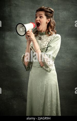 Vintage portrait of young shouting girl in medieval style dress with megaphone isolated on dark background. Comparison of eras concept. News, sales Stock Photo