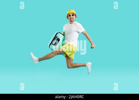 Joyfully excited guy in summer clothes runs and jumps with suitcase on light blue background. Stock Photo