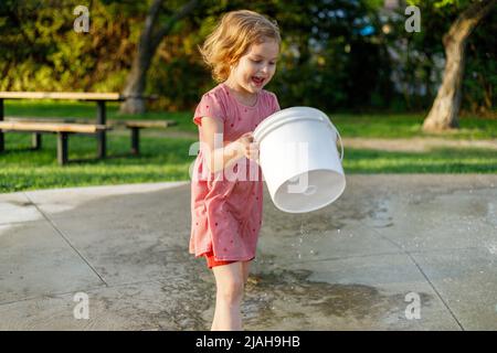 Happy smiling child playing with bucket at splash pad in park in summer. Little kid outdoors Stock Photo