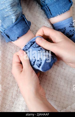 A mother woman wears blue socks to a foot baby boy. Mom dresses a leg child in clothes on the bed. Stock Photo