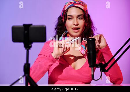 Self-confident attractive young woman sitting in front of smartphone camera creating content for ASMR blog recording sound of lipstick opening Stock Photo