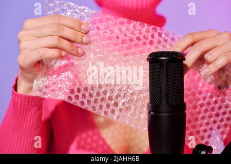 Unrecognizable young female influencer wearing pink clothes popping bubble wrap on microphone for ASMR content Stock Photo