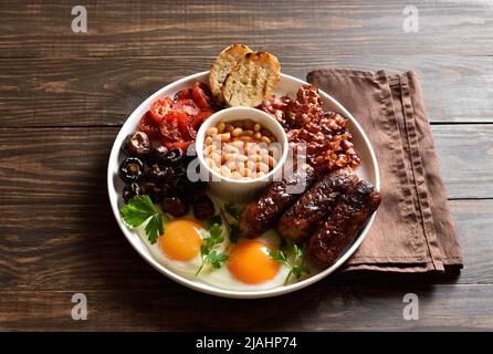 Full English breakfast with fried egg, bacon, sausage, beans and mushrooms on wooden background. Stock Photo