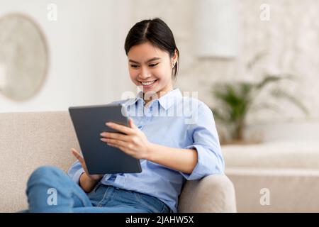 Smiling lady sitting on sofa, using digital tablet Stock Photo