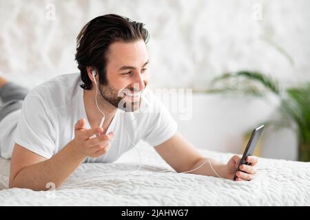 Joyful guy laying on bed, using smartphone and earphones Stock Photo