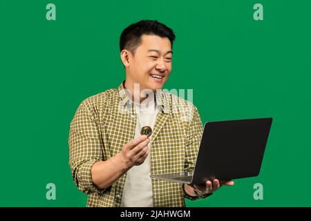 Happy mature asian man holding golden bitcoin and laptop, earning cryptocurrency, standing over green background Stock Photo