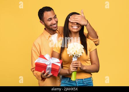 Handsome young black man giving flowers and gift box to his girlfriend, covering her eyes, making birthday surprise Stock Photo