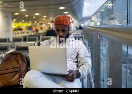 Young hipster black man sitting in airport terminal using laptop wear headphones and listening music Stock Photo