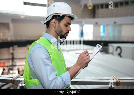 Thoughtful millennial islamic man engineer in protective uniform and hard hat with beard makes notes in tablet Stock Photo