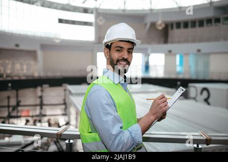 Glad young islamic professional man engineer builder with beard in protective clothing and hard hat makes notes Stock Photo