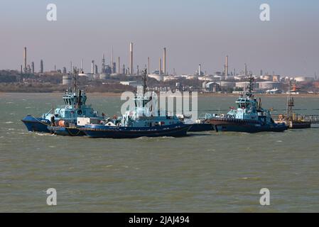 Fawley, Southampton, England, UK. 2022.  Three ocean going tugs alongside a jetty with a background of Fawley Refinery, storage tanks, chimneys and sm Stock Photo