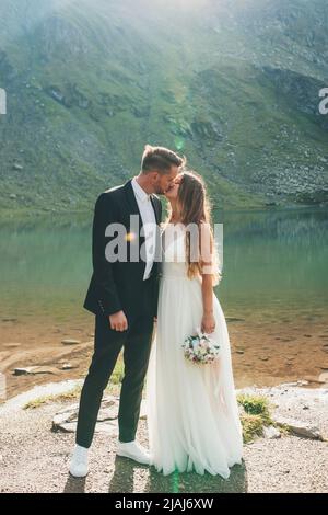 Newlyweds in love kissing standing against the backdrop of lake and mountains landscape. Outdoor landscape. Beautiful summer landscape. Nature Stock Photo