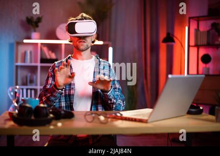 Young caucasian man sitting at home office and using VR glasses and laptop for remote work. Attractive male freelancer touching virtual screen with fingers. Stock Photo