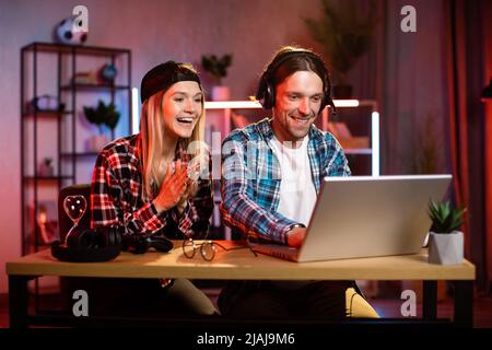 Overjoyed blond woman in cap and her young man in headset gesturing and screaming while looking at screen of laptop. Young caucasian couple celebrating win of favorite team. Stock Photo