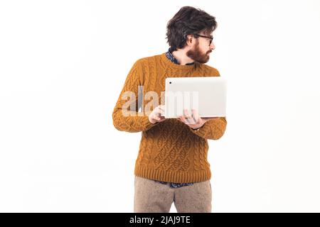 Young caucasian bearded entrepreneur in a knitted sweater standing in a studio, holding his thin silver laptop, and looking back at white background. High quality photo Stock Photo