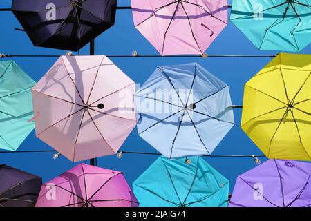 Umbrellas hanging from cables creating a ceiling effect; rows of decorative pastel colored parasols made translucent in the bright sun filled blue sky Stock Photo