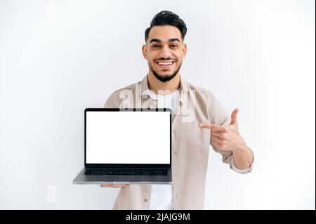 Joyful arabian or indian guy in casual wear, freelancer or student, holding an open laptop with empty mock-up space and points finger on it, stand on white isolated background, looks at camera, smiles Stock Photo