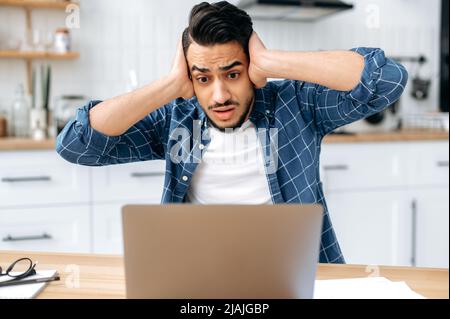 Frustrated sad puzzled arabian or indian guy, freelancer or designer, got a negative result, failed the project, looks desperately at laptop screen, holds his hands on a head, have stressed situation Stock Photo