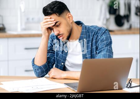 Overworked from work, deadline. Sad arabian or indian guy, sitting at a desk, looking tired and stressed, frustrated looking at work graphs, failing to finish work on the project on time, needs rest Stock Photo
