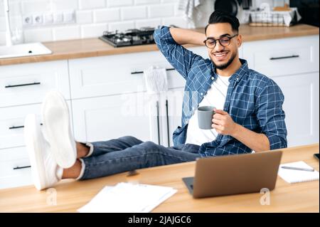 Chilling during work. Positive indian or arabian guy in stylish clothes, sit at home in the kitchen with his legs on the table, holding a cup of coffee in hand, working remotely from home using laptop Stock Photo