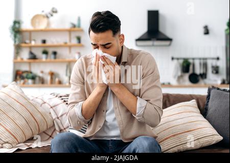 Unhappy sad indian or arabian guy suffering from fever and flu, sitting on sofa in living room, blowing nose and sneezing in napkin, need treatment of illness, and doctors consultation Stock Photo